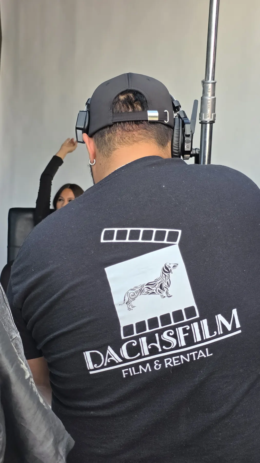Operador de cámara de espaldas, con polera DACHSFILM, encuadrando a la talento.